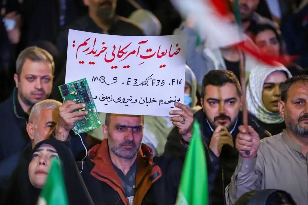 Supporters attend a pro-government gathering in Iran, where one man holds a placard reading “We buy American scraps,” alongside references to US military equipment including F-16 and F-35 fighter jets, MQ-9 drones and naval destroyers.