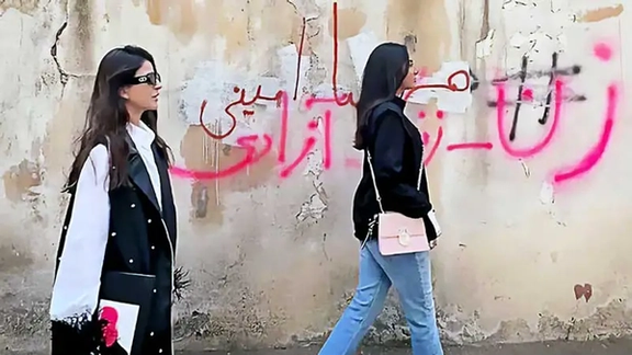 Women walking on the streets of Iran without mandatory hijab. The slogan on the wall says: "Woman, Life, Freedom"