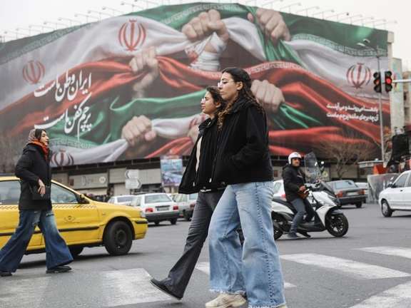Pedestrians cross a street in Tehran with a large mural depicting clenched fists holding the Iranian flag looming in the background, January 19, 2026