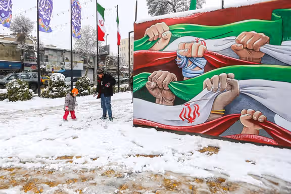 A toddler and a young boy play in the snow next to a state-sponsored billboard depicting Iranian fists and flags, in northern city of Qazvin, January 20, 2026