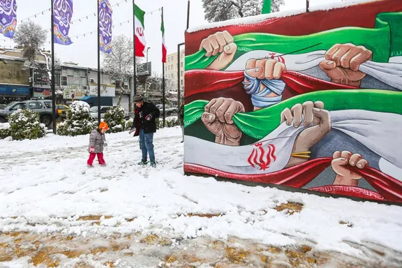 A toddler and a young boy play in the snow next to a state-sponsored billboard depicting Iranian fists and flags, in northern city of Qazvin, January 20, 2026