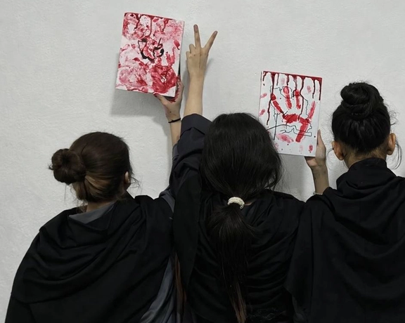 Three young women hold up red-painted artwork representing the blood of innocent protestors, including handprints and Persian script, while one raises a victory sign.