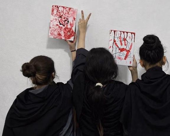 Three young women hold up red-painted artwork representing the blood of innocent protestors, including handprints and Persian script, while one raises a victory sign.