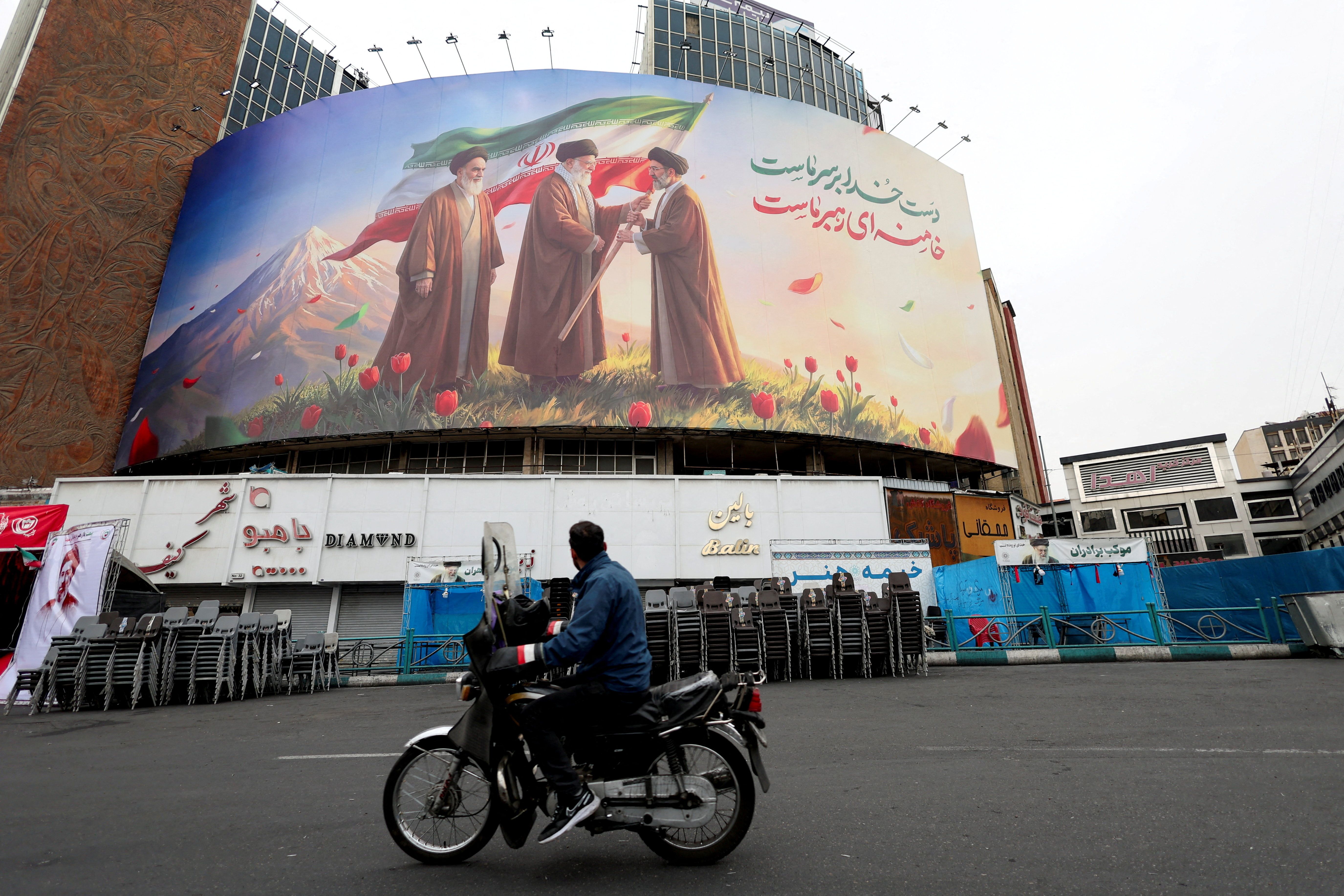 A man on a motorcycle looks at a large billboard featuring Iran's new Supreme Leader Mojtaba Khamenei, and late Supreme Leaders Ali Khamenei and Ruhollah Khomeini, amid the US-Israeli conflict with Iran, in Tehran, Iran, March 12, 2026. 