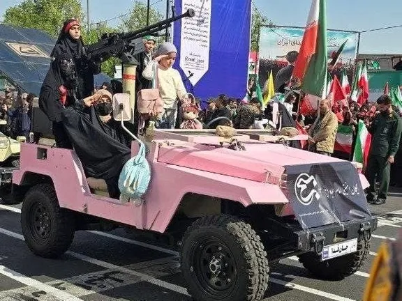 A pink military-style vehicle moves through a street rally in Iran, carrying several women dressed in black chadors and hijabs.