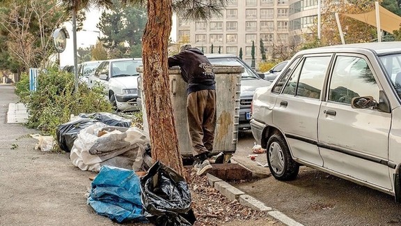 A man searches through a garbage bin on a city street in Iran, highlighting the harsh realities of poverty and economic struggle faced by many residents.