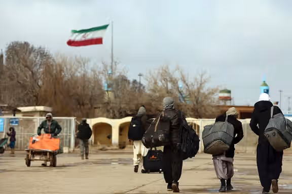 Afghan migrants walk away with their belongings as the Iranian flag flies in the background