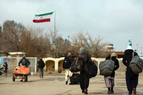 Afghan migrants walk away with their belongings as the Iranian flag flies in the background