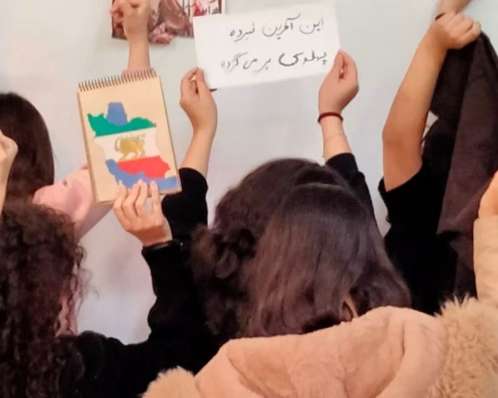 Schoolgirls in Iran raise fists in protest; a handwritten sign reads, “This is the final battle, Pahlavi will return.”