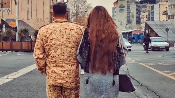 A woman defying Iran's hijab regulations walks alongside a conscript in Tehran's Valiasr crossroad in December 2024