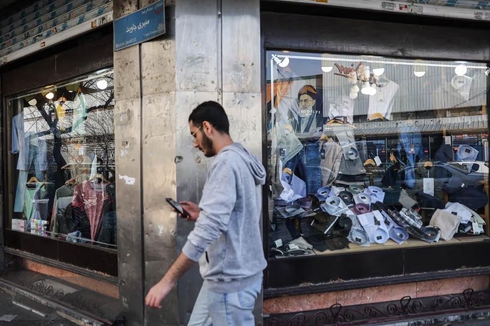 A young man looks at his mobile passes by a retail shop with a picture of Iran's slain supreme leader Ali Khamenei on display, Tehran, Feb. 12, 2026