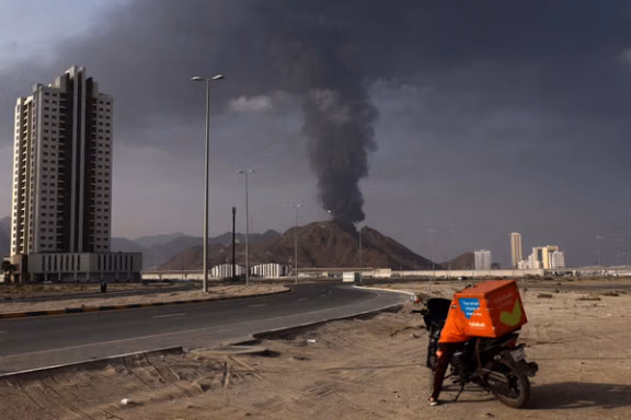 A person stands next to a motorcycle as smoke rises in the Fujairah oil industry zone following a fire caused by debris after interception of a drone by air defenses, according to the Fujairah media office, United Arab Emirates, March 3, 2026.