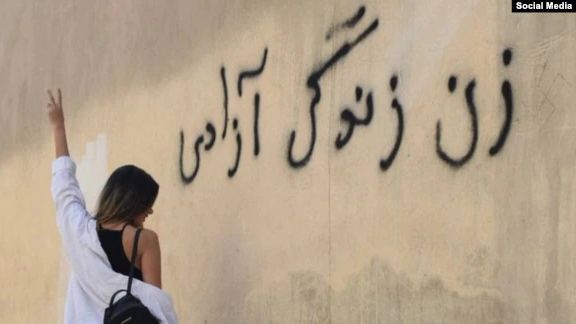 An Iranian woman standing in front of a wall on which the main slogan of the ongoing protests – Women, Life, Liberty – is seen