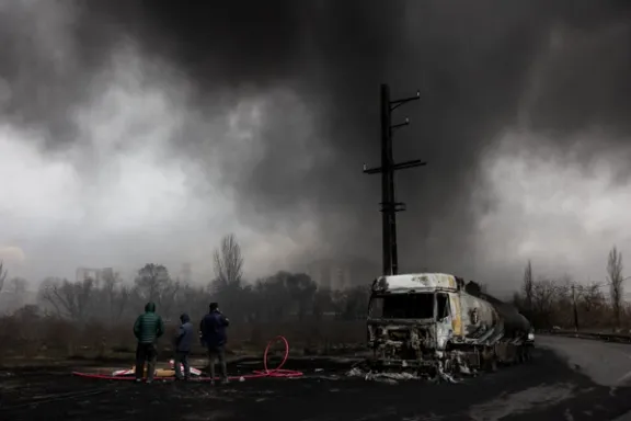 People stand near a destroyed vehicle as smoke rises after a reported strike on Shahran fuel tanks, amid the US-Israeli conflict with Iran, as seen through a window, in Tehran, Iran, March 8, 2026.