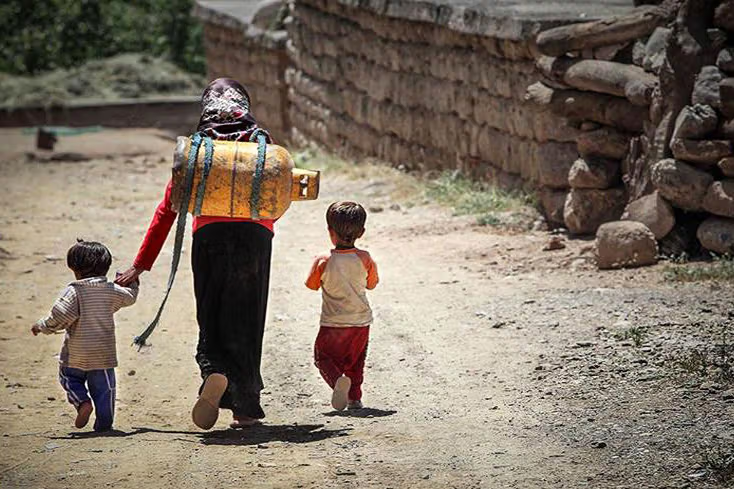 A woman carries a gas capsule on her back as she walks with two children along a dirt road in a rural area of Iran.